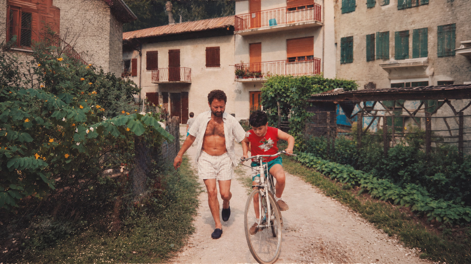 A man in an open shirt walks beside a boy on a white bicycle along a path between houses: the photo on the cover of Old Man by colourshop